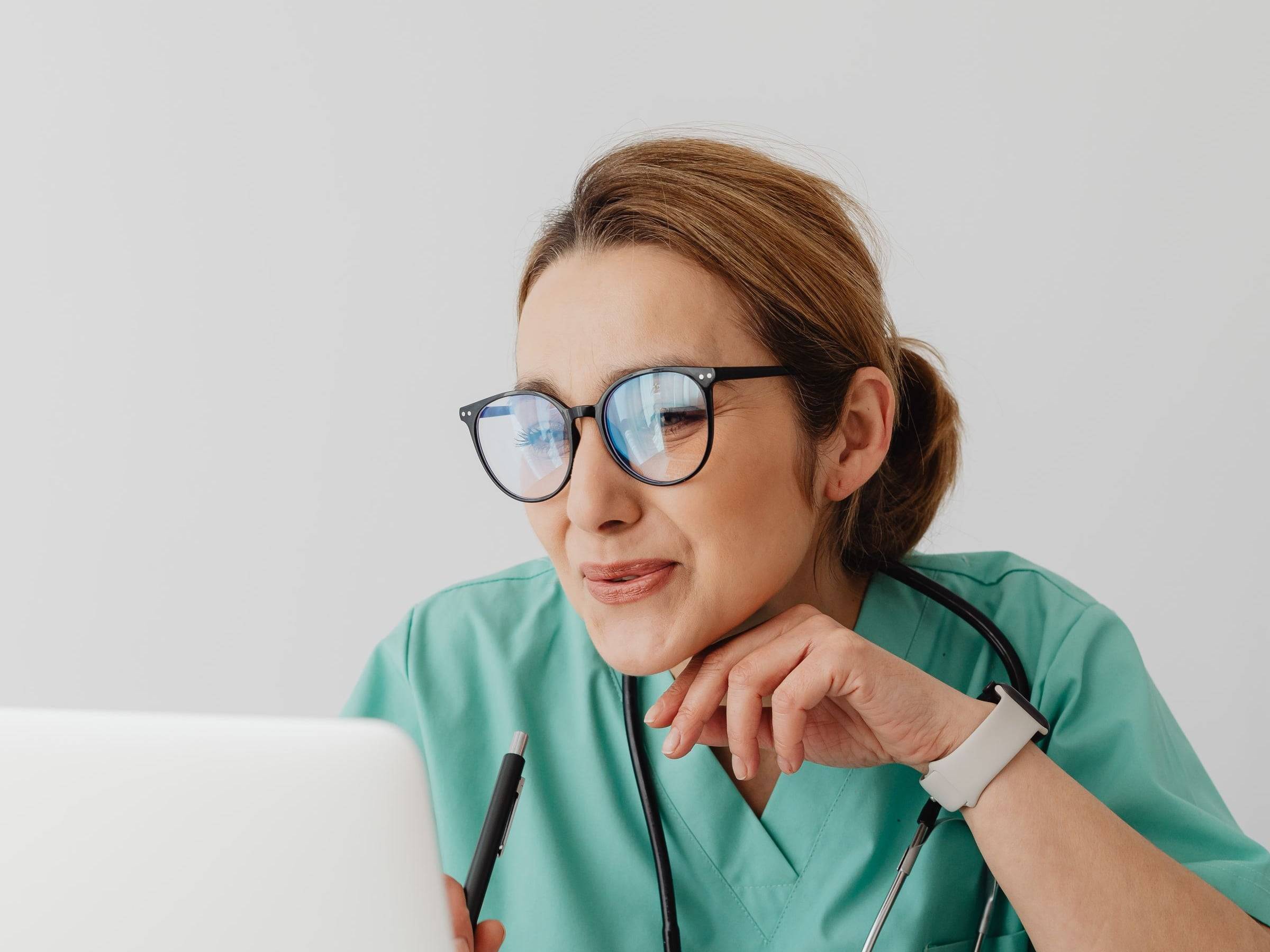 A clinician in scrubs, with a stethoscope around her neck, works at a laptop.