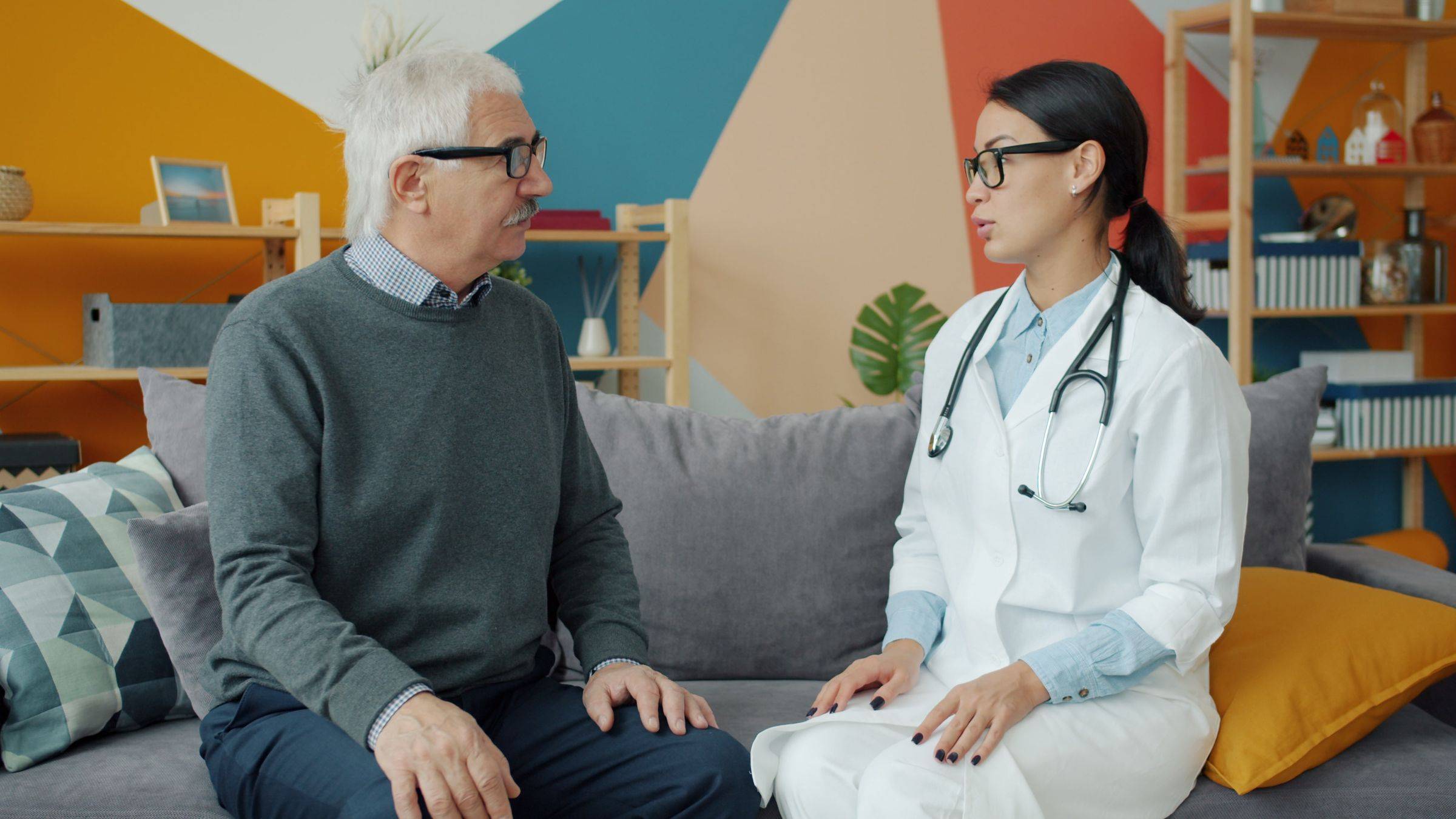 A clinician in a white coat talks with an older man in a bright living room.